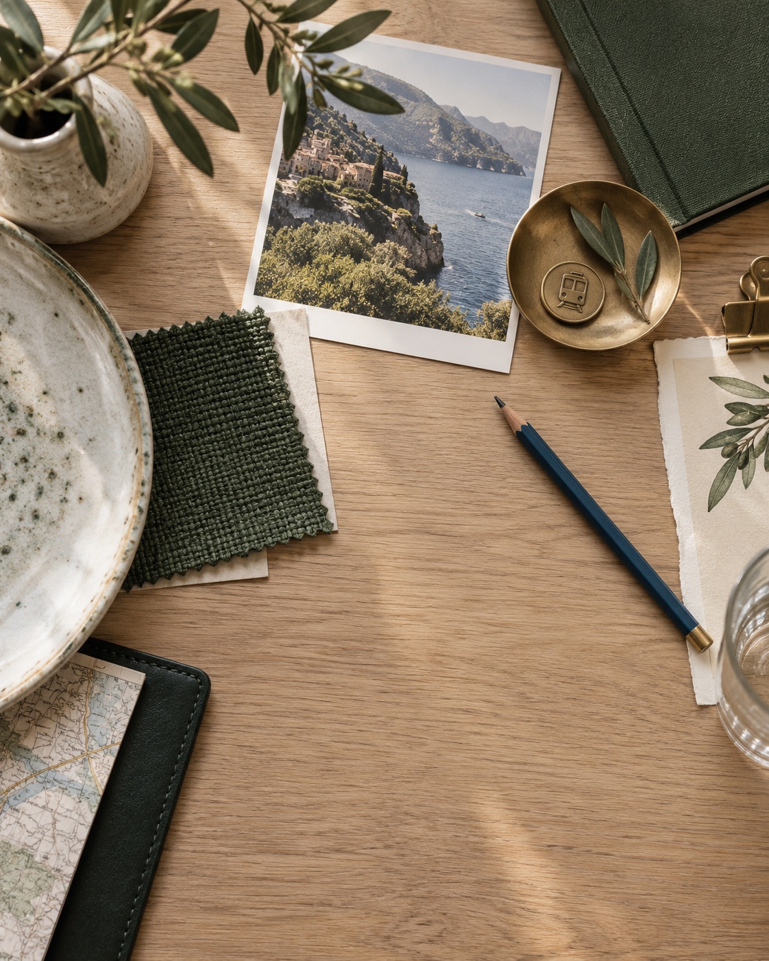 A pale oak table with a coastal photograph, a fabric swatch, a brass dish and a pencil